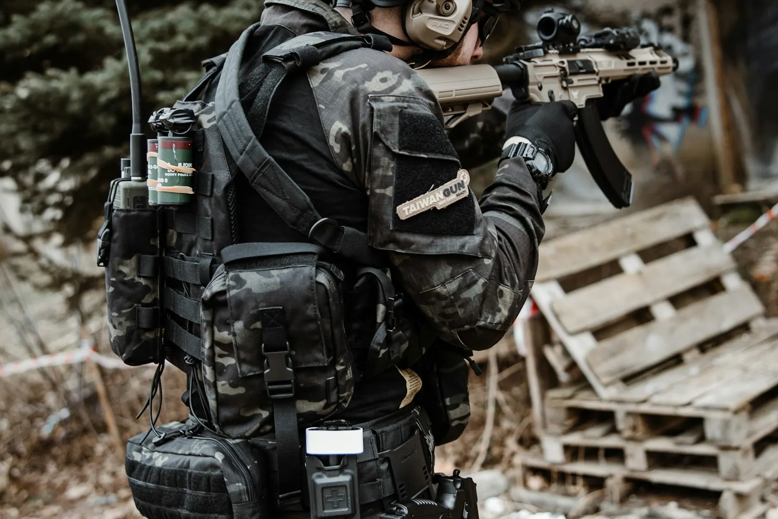 Joueur d'airsoft en tenue noire et camouflage visant près d'un tas de palettes en bois sur un terrain aménagé