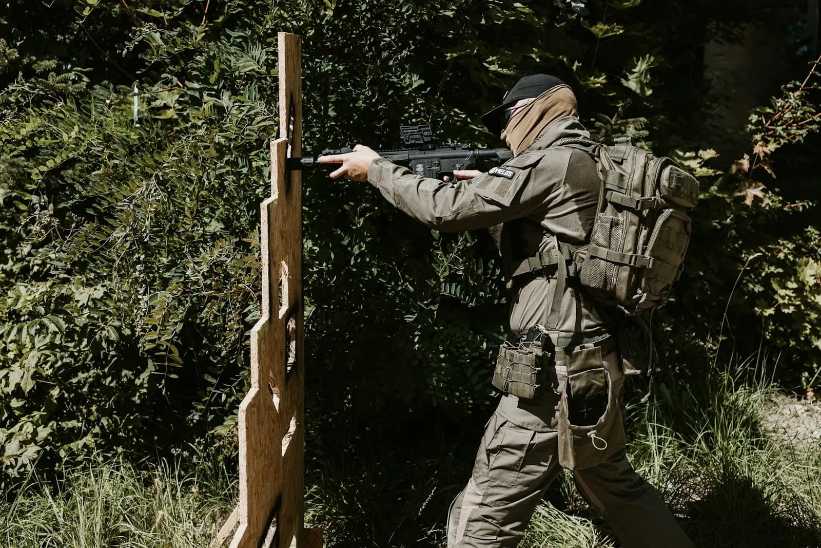 Joueur d'airsoft en tenue beige avec sac à dos, visant derrière une barricade en bois dans un sous-bois