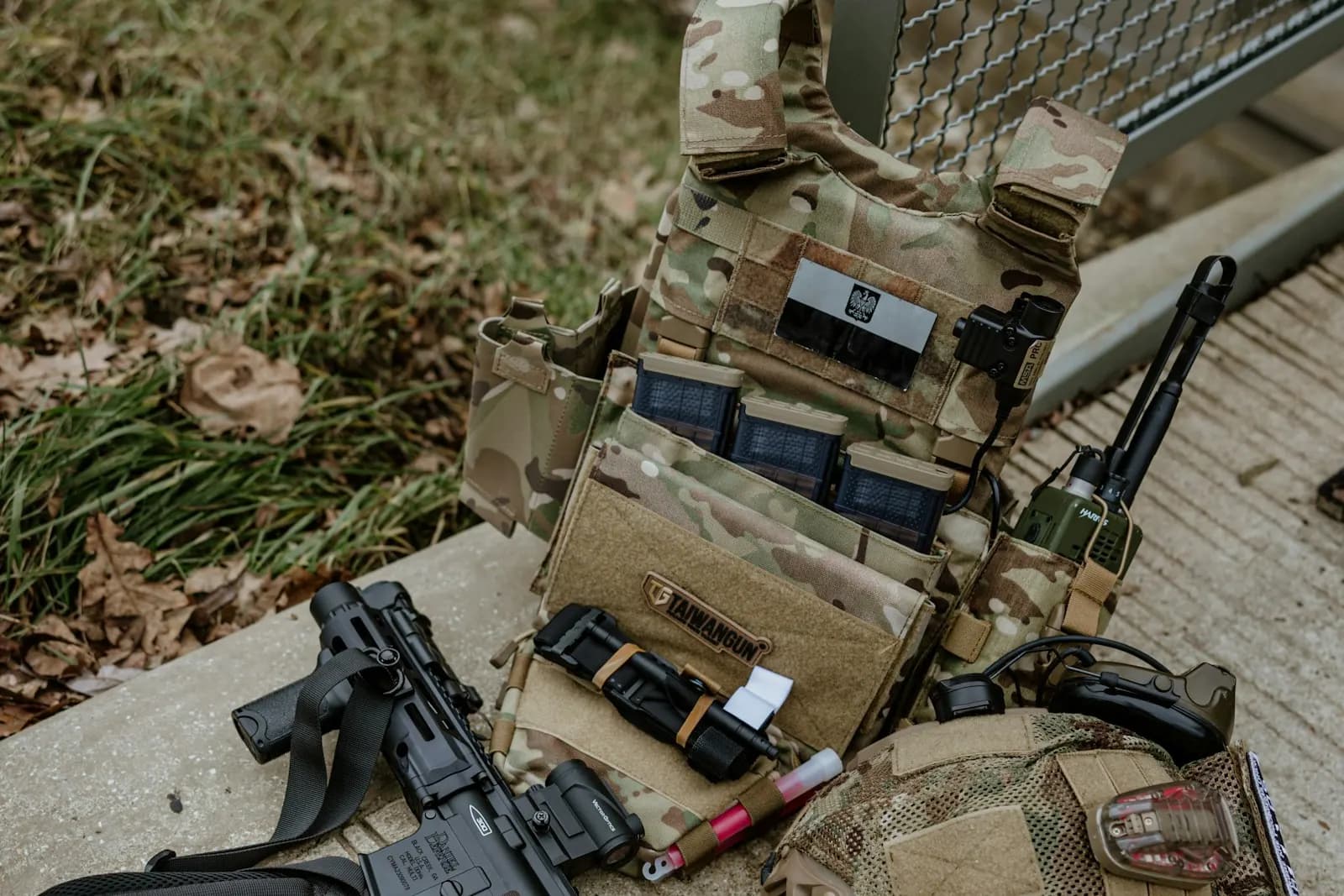 Équipement d'airsoft posé au sol : réplique AEG, gilet porte-plaques avec chargeurs, casque et radio