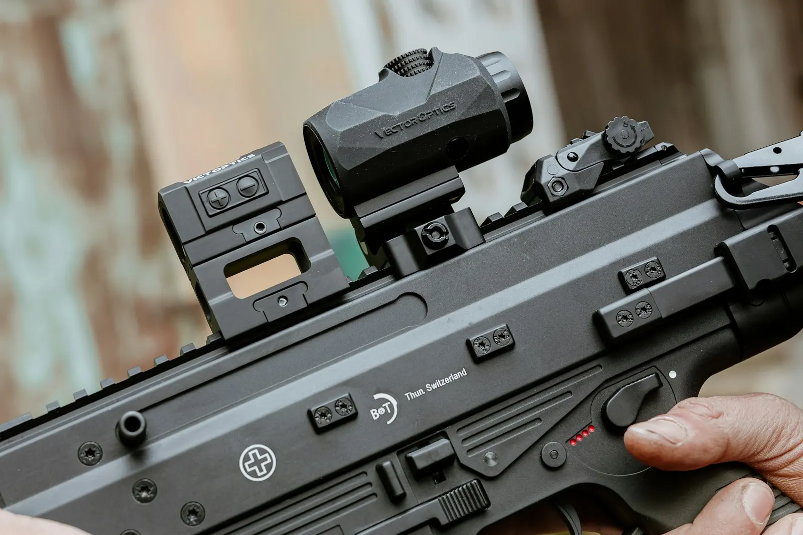 Gros plan sur une réplique d'airsoft noire équipée d'un point rouge et d'un magnifieur, main sur le sélecteur de tir