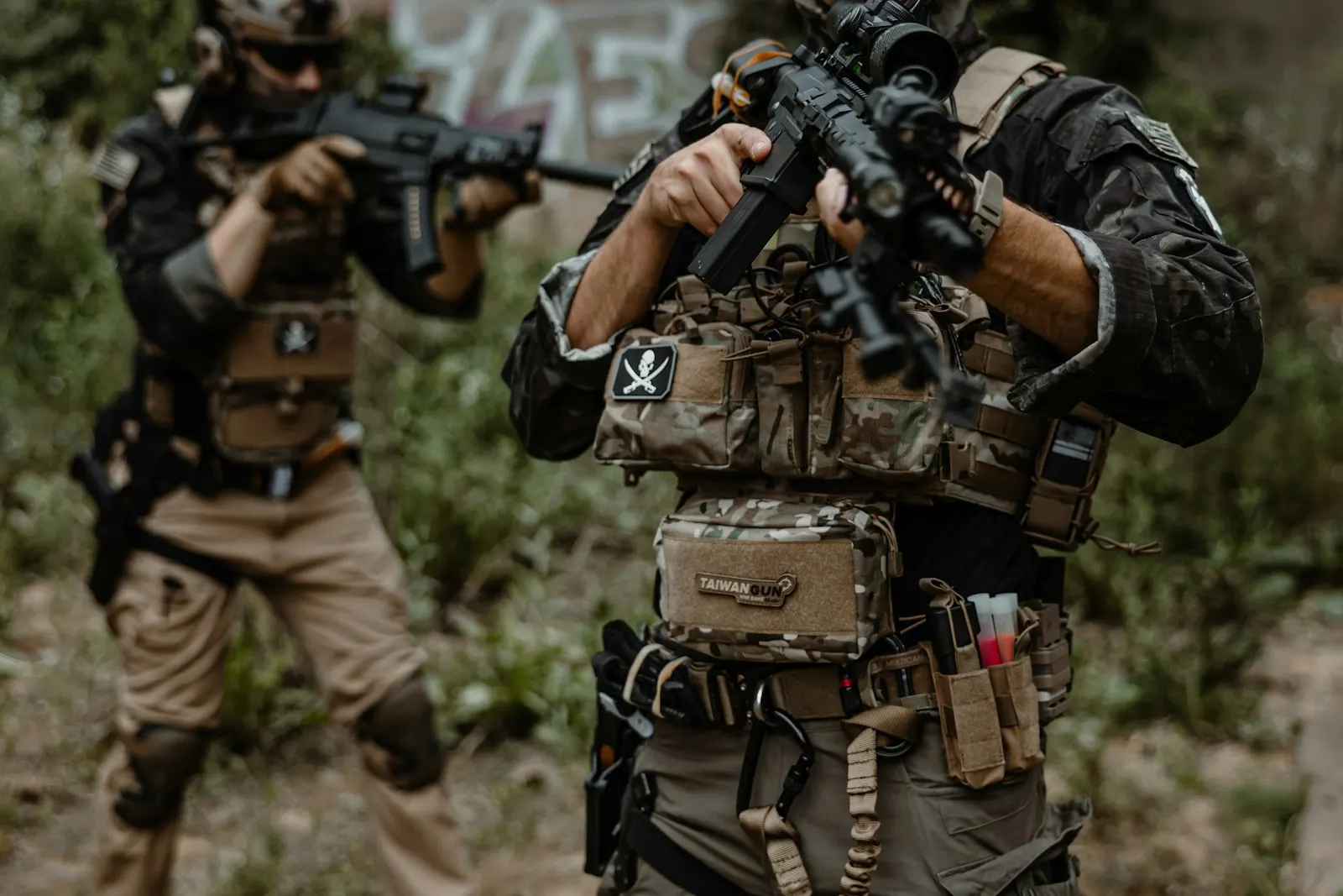 Deux joueurs d'airsoft en tenue tactique complète progressant en binôme, répliques épaulées, dans une zone boisée