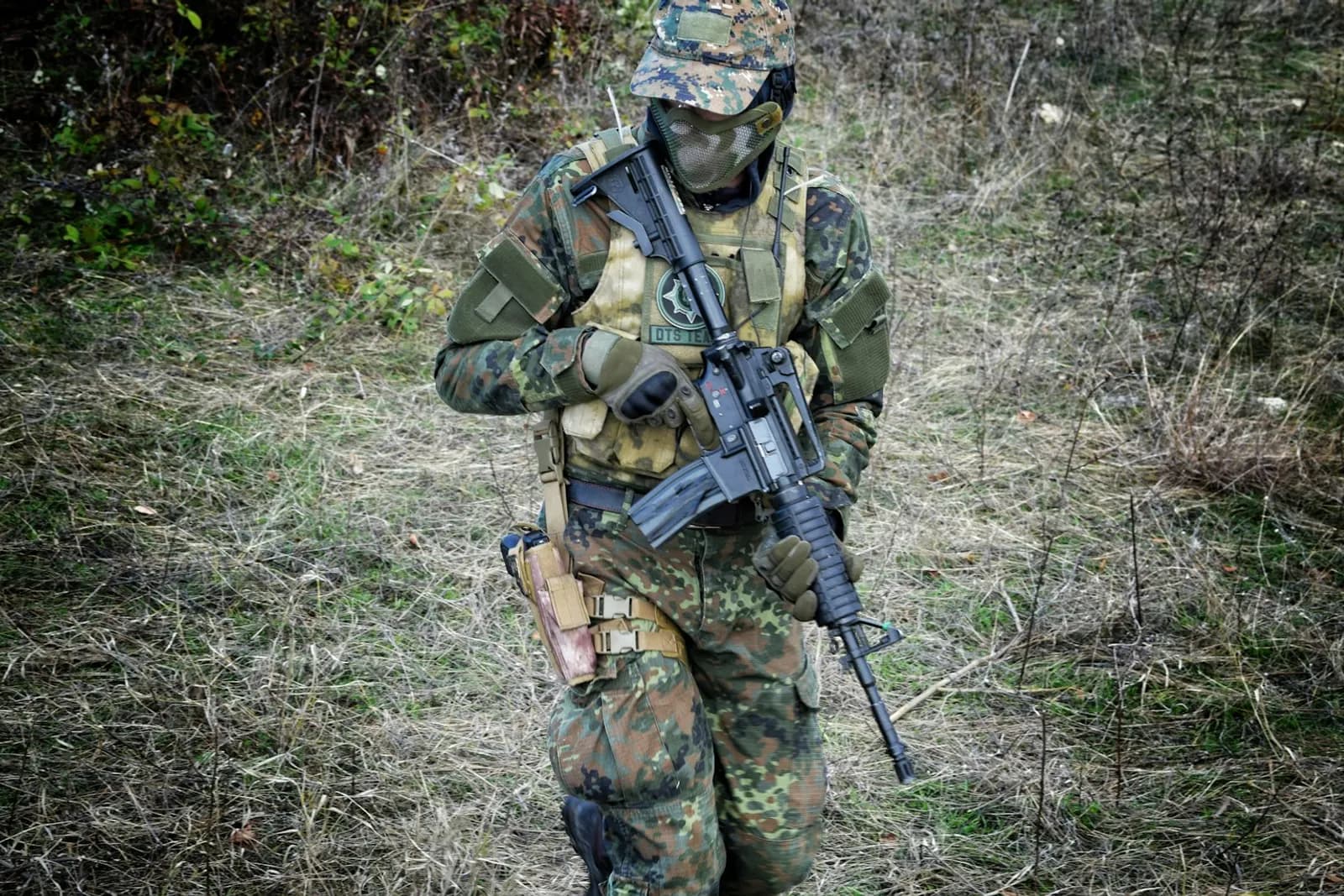 Joueur d'airsoft en tenue camouflage complète avec casque, gilet tactique et réplique, marchant dans les herbes hautes