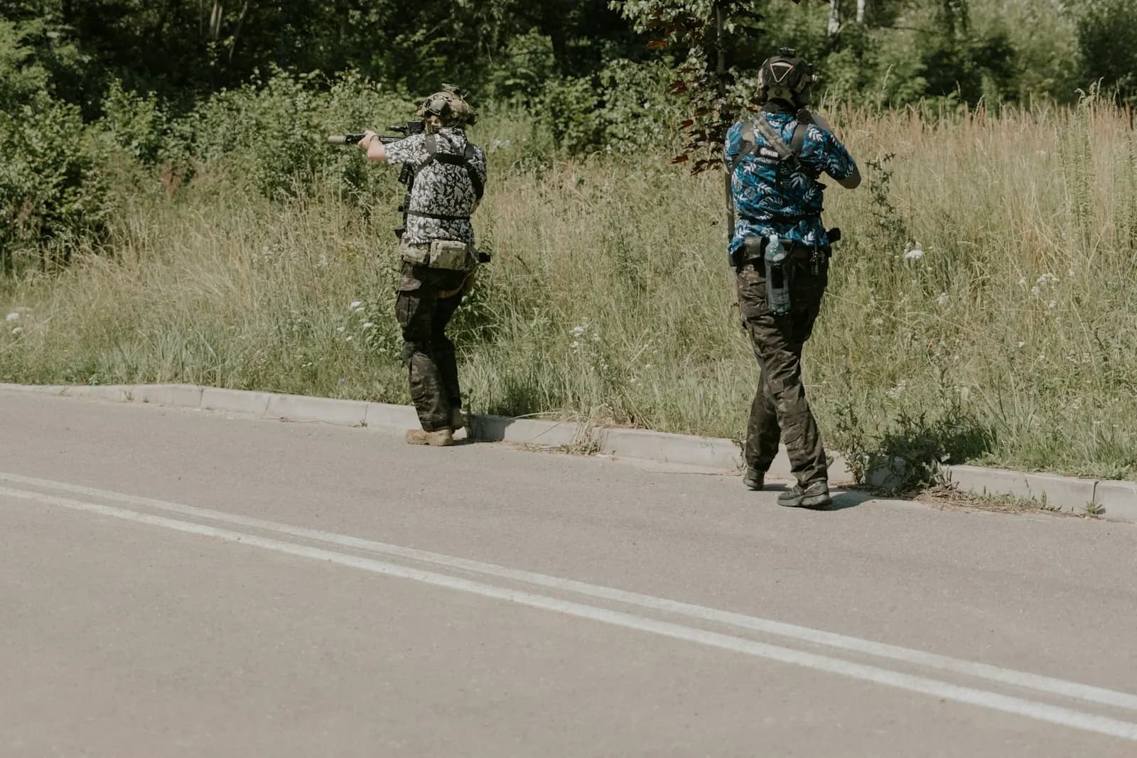 Deux joueurs d'airsoft progressant le long d'une route en lisière de forêt, répliques épaulées